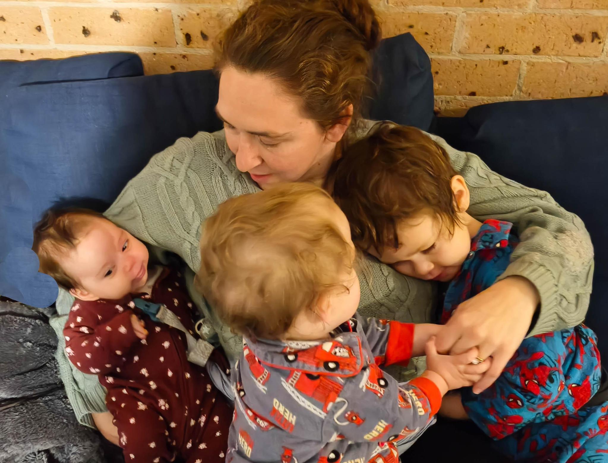 Vivienne sitting on a couch cuddling her three young children in pajamas, with an exposed brick wall behind them
